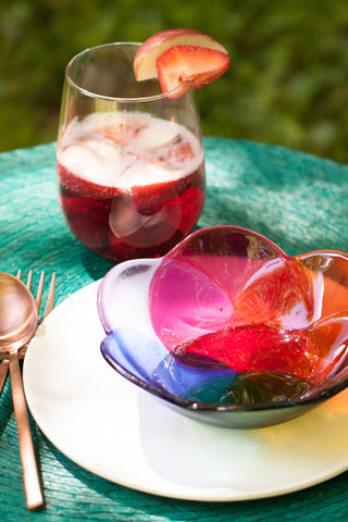 handmade Fused glass bowl in a floral shape in rainbow colors made by people with disabilities shown with a teal placemat and glass of wine