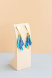 Plume drop resin earrings in blue and teal with gold triangular findings hanging to show the layered teardrop effect